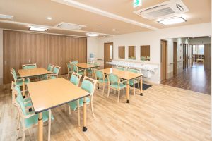 介護施設の広々とした活動室。木製テーブルと緑色の椅子が配置され、レクリエーションや交流に使用される共用スペース。